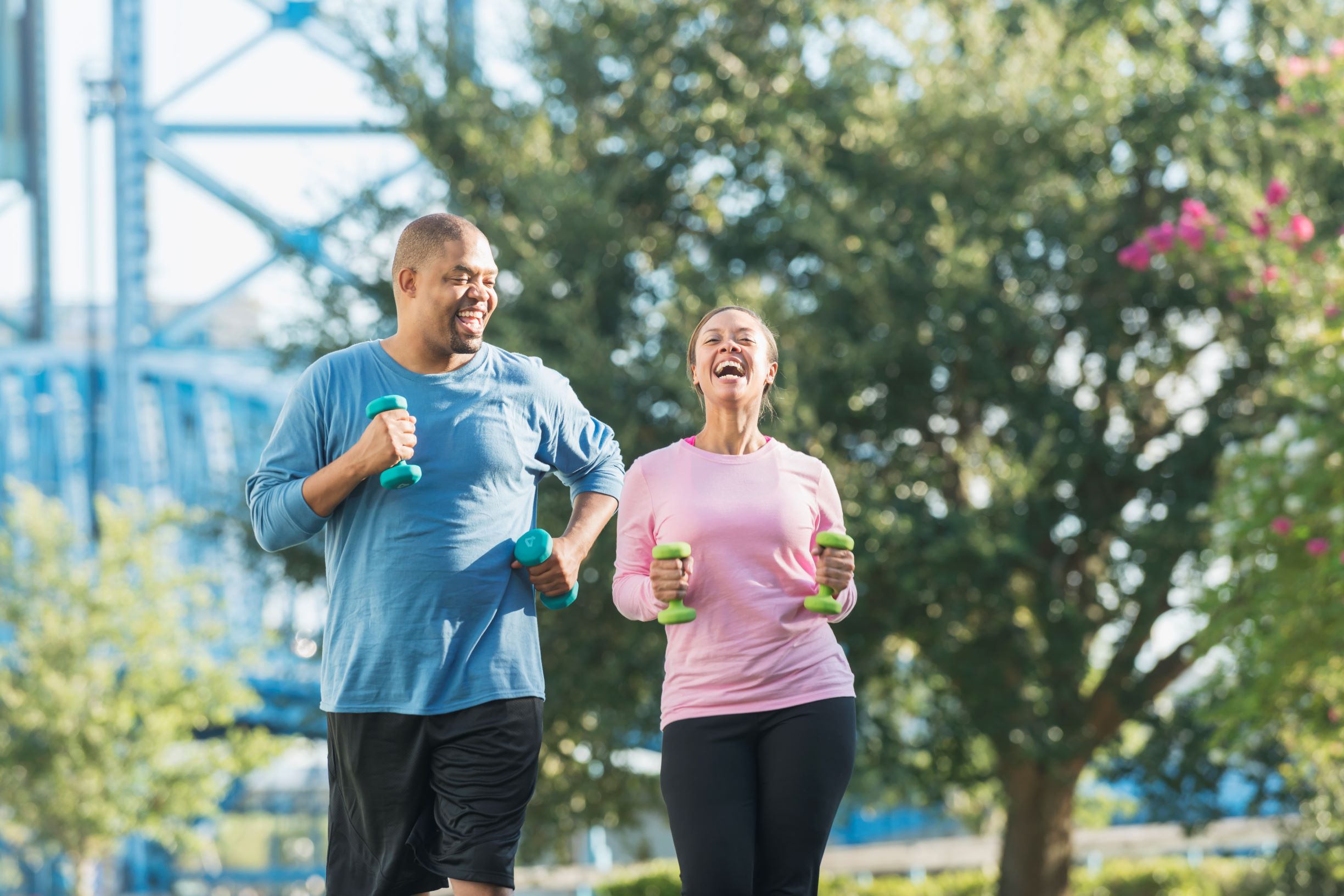 A couple walk with dumbbells in each hand while laughing. Carrying dumbbells during your walk gives you the added benefit of building up those arm muscles. Having someone you love walking with you as well makes it all that much better.