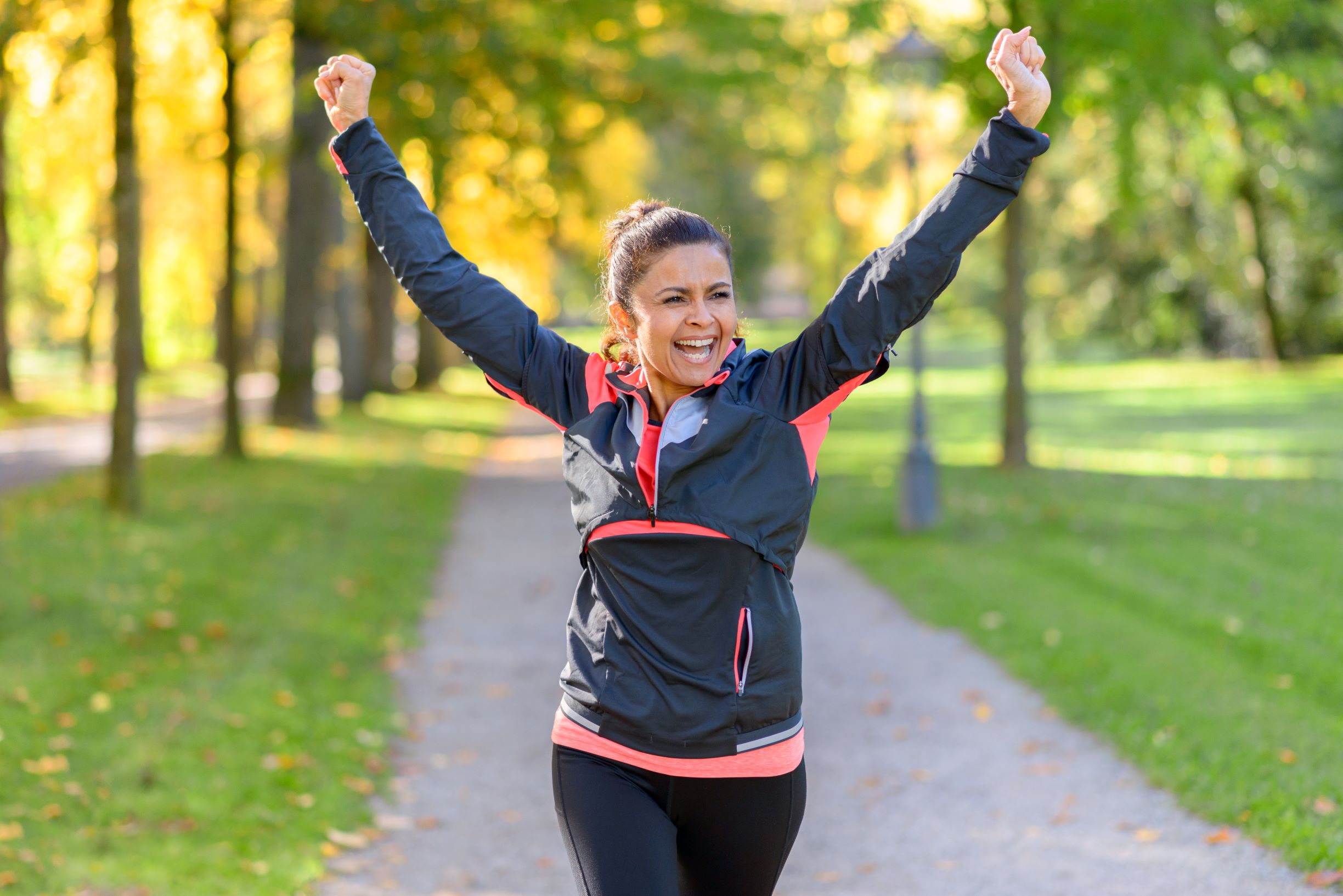 There are many fun ways to walk for heart health. In this image, a woman is walking in the park while raising her arms in celebration.