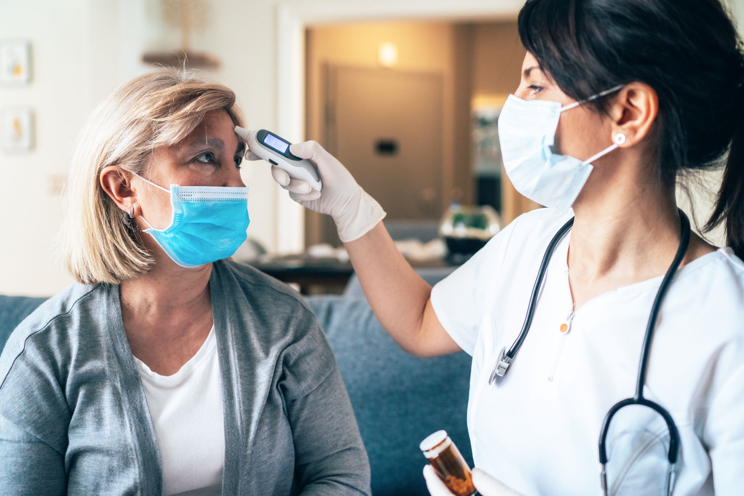 Obesity increases the risk for COVID-19 complications for various reasons, as obesity itself opens people up to many health conditions. In this image, a medical provider takes a patient's temperature. Both are wearing masks over their nose and mouth.